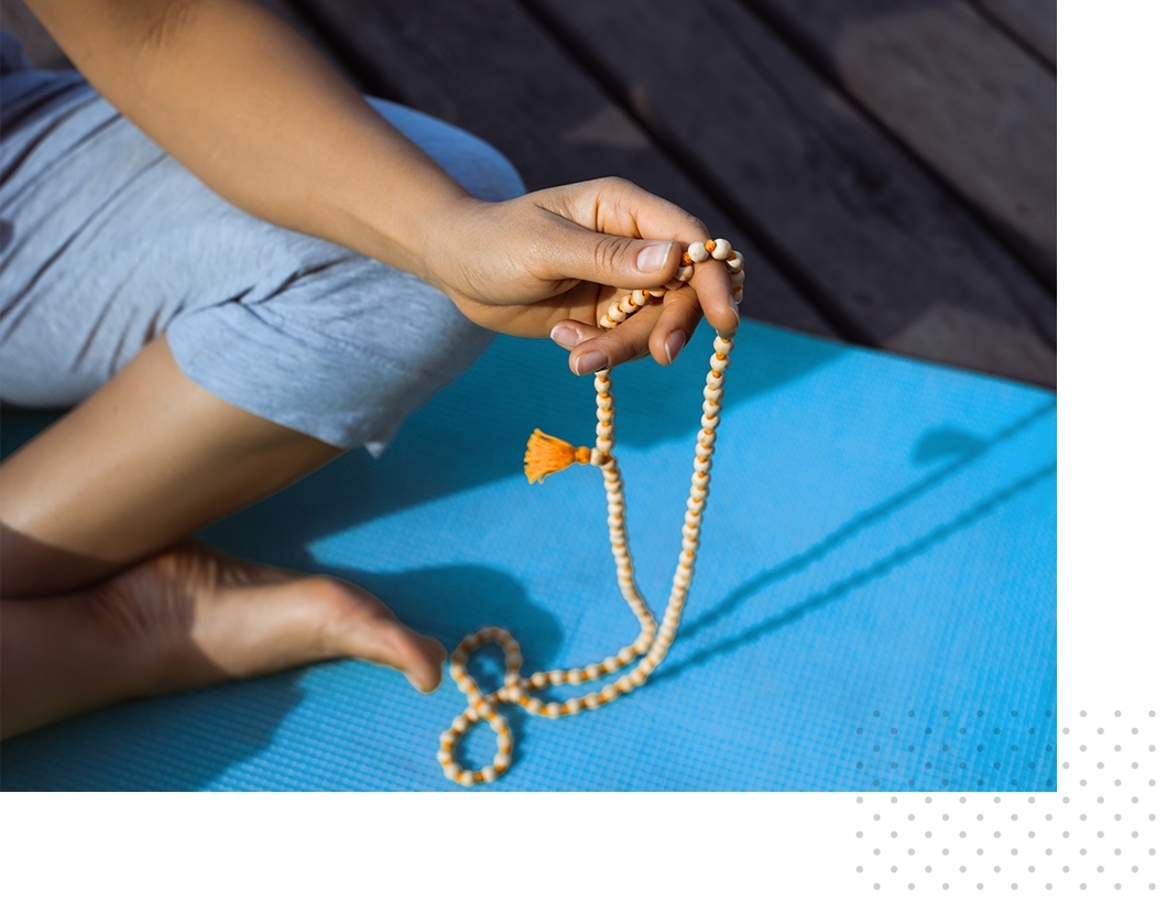 An individual sitting crossed-legged and holding a beaded necklace in one hand.