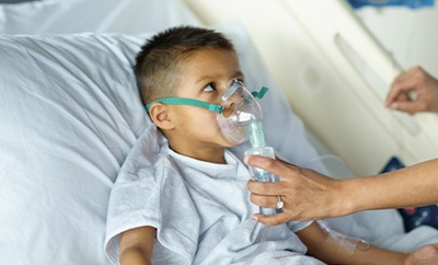 A hispanic child in a medical bed uses a respirator with support from a medical professional.