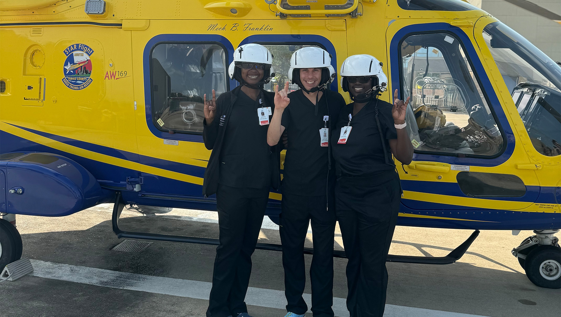 Medical trainees in black scrubs gather in front of an emergency medicince helicopter.