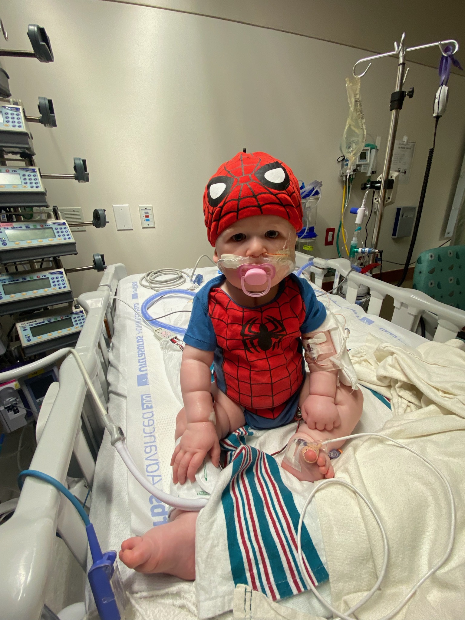 An infant wearing a Spiderman outfit, and affixed with medical tubes and apparatuses, sits on a medical gurney.