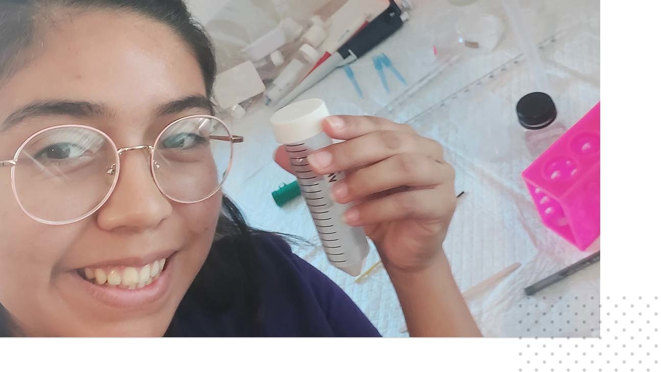 Valeria, a 2022 SHE participant holds a test tube, with varying paperwork and science equipment in the background.