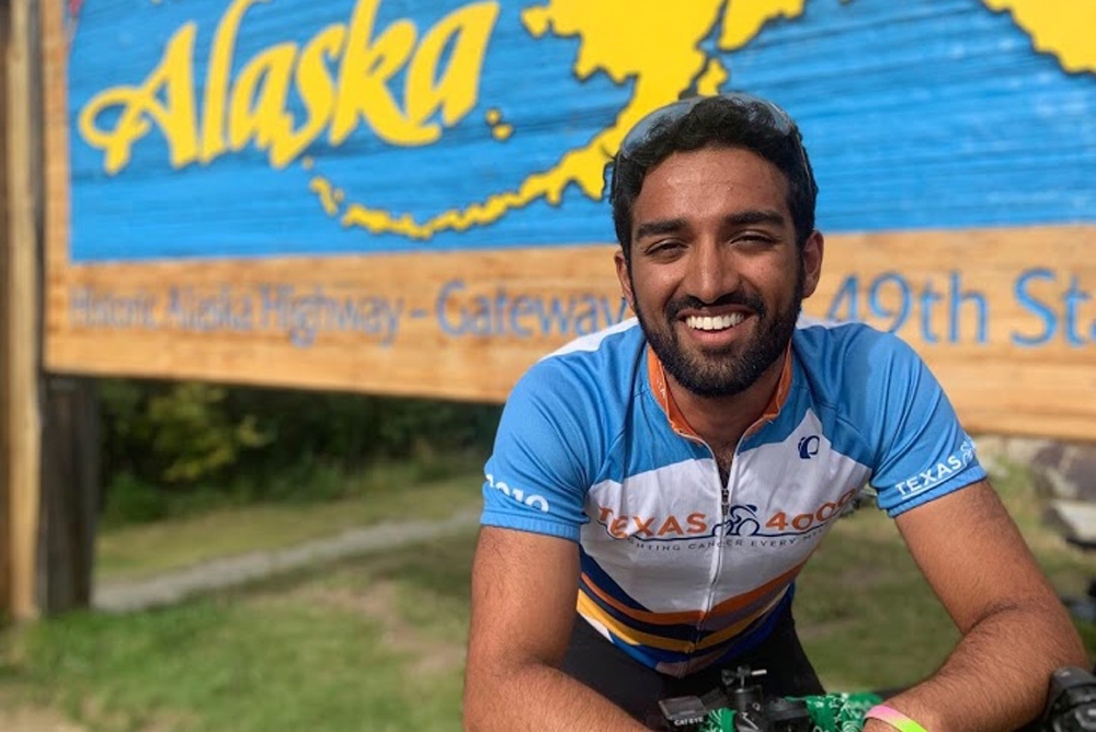 Shiva Velingker leaning on a bicycle in front of the welcome to Alaska sign.