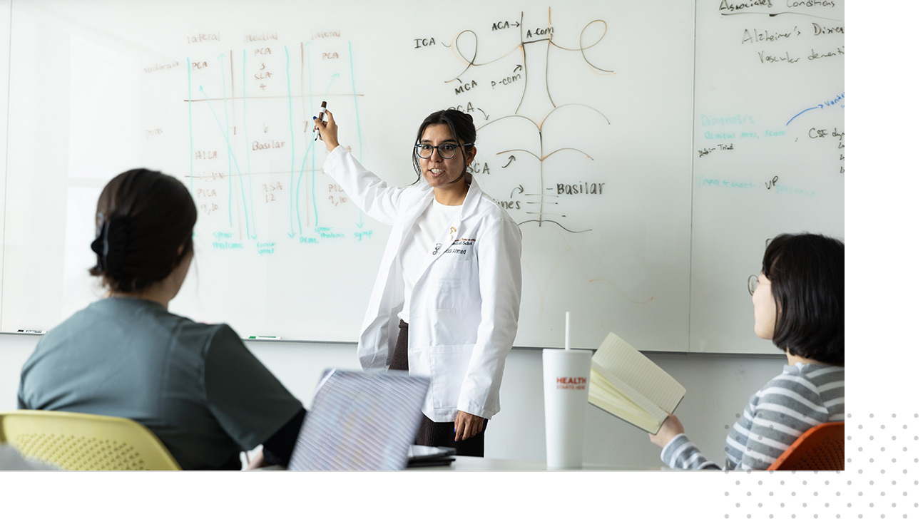 A Dell Medical School student wearing a white coat points to a whiteboard and discusses the content written on it to other students.