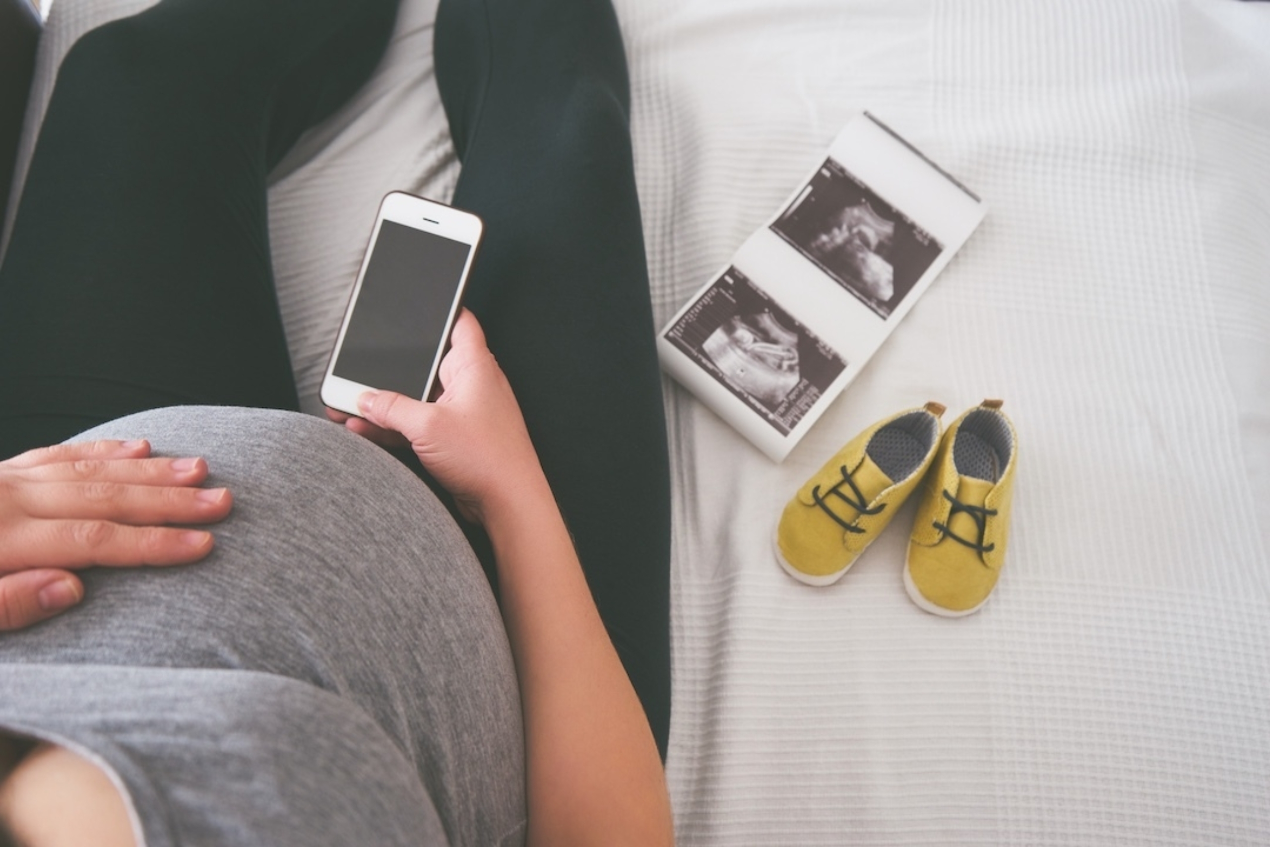 Image: An overhead view of a pregnant woman's hand on her stomach, holding a smartphone. Next to her are ultrasound scans and baby shoes.