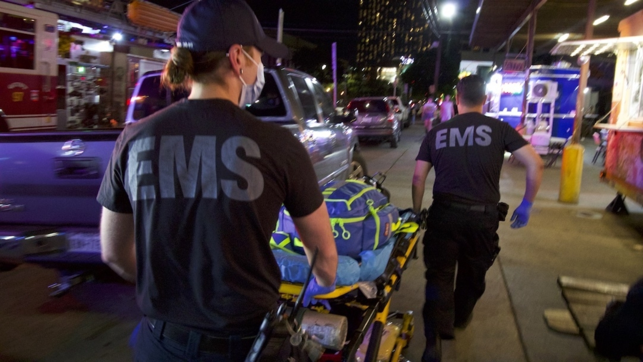 Image: Two paramedics carry a stretcher away from the camera on a city street.