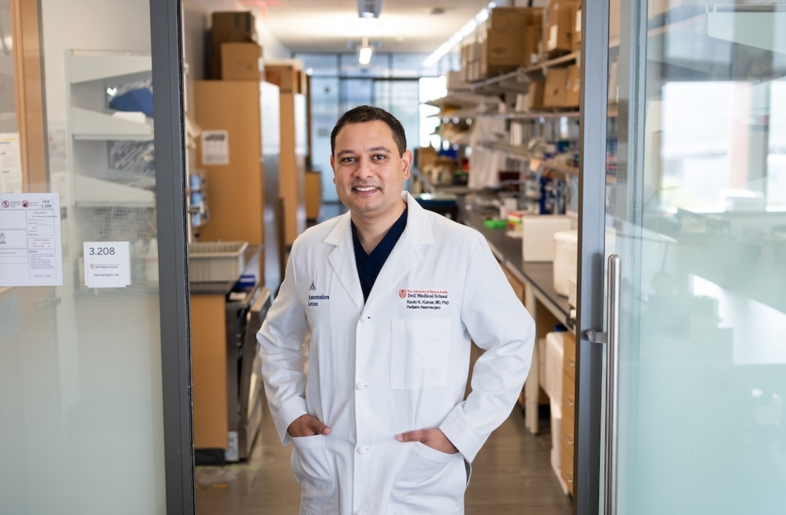 Kevin Kumar stands in white coat, hands in his pockets, looking at the camera from the doorway of his research lab.