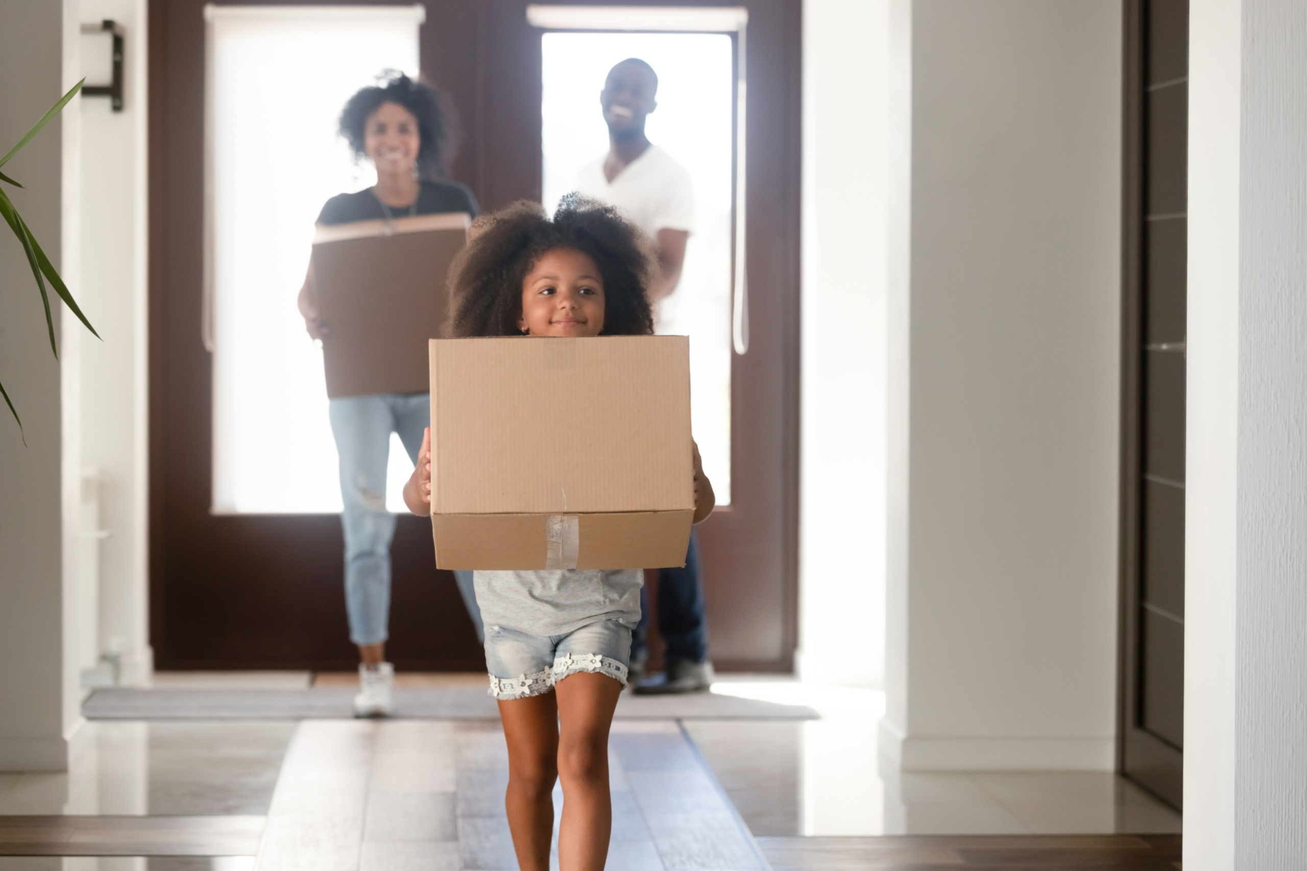 Little girl carries moving box into home, while her parents follow behind.