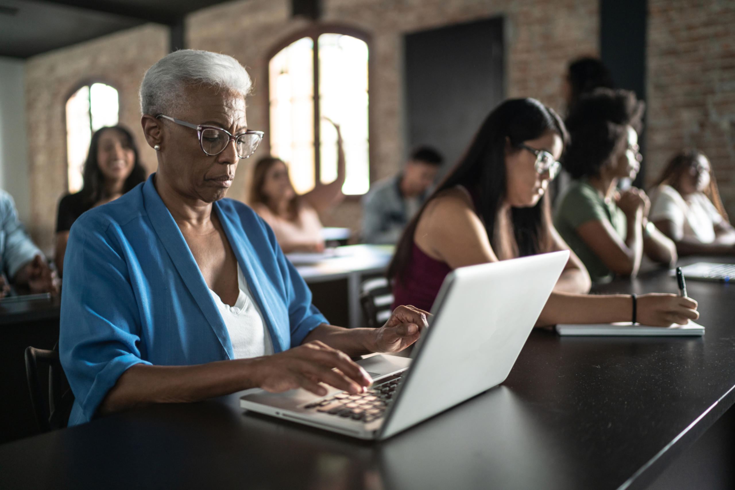 Older woman uses laptop while sitting in a classroom environment.
