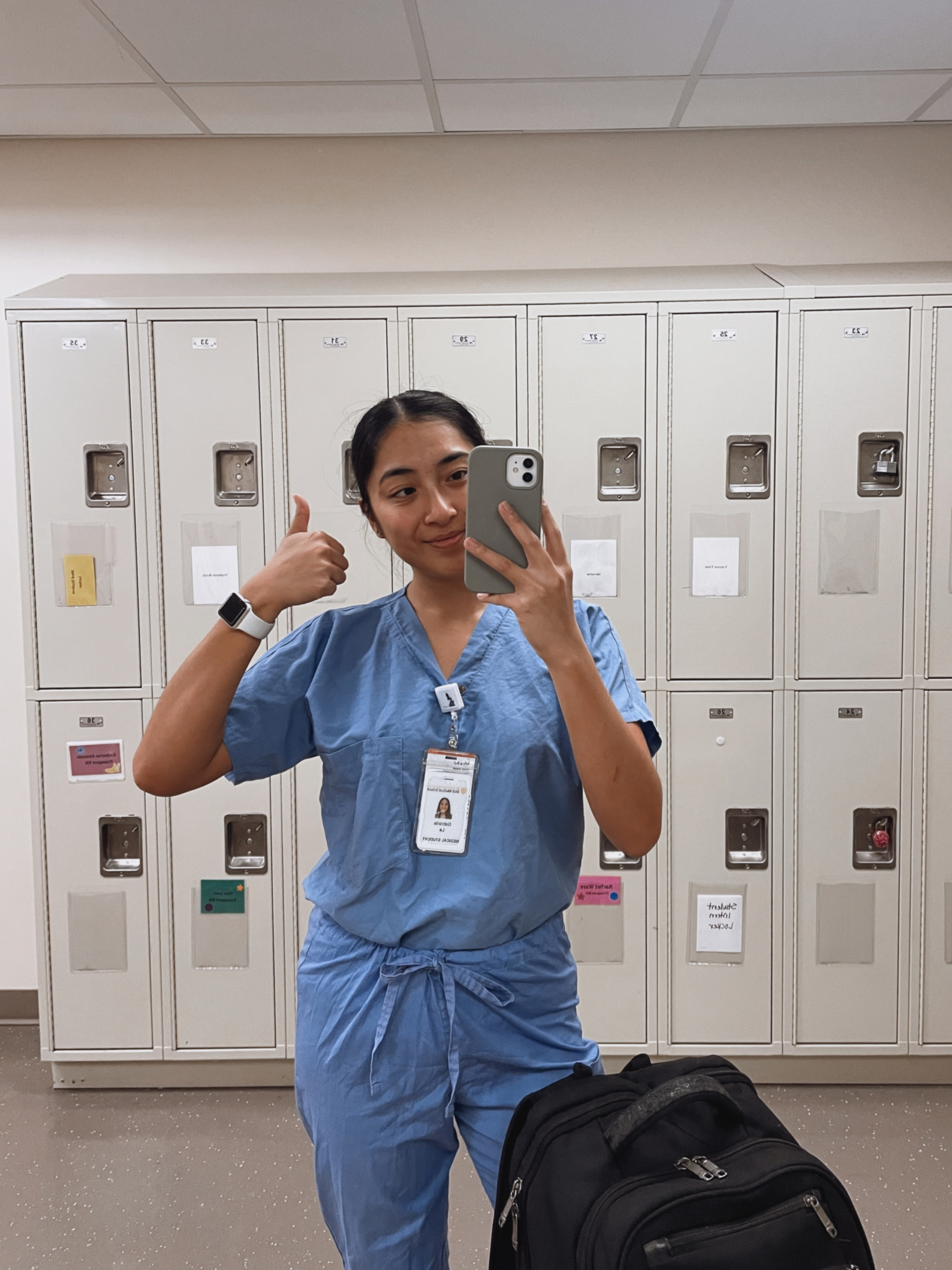 A Dell Medical School student wearing scrubs takes a mirror selfie in front of a row of lockers.
