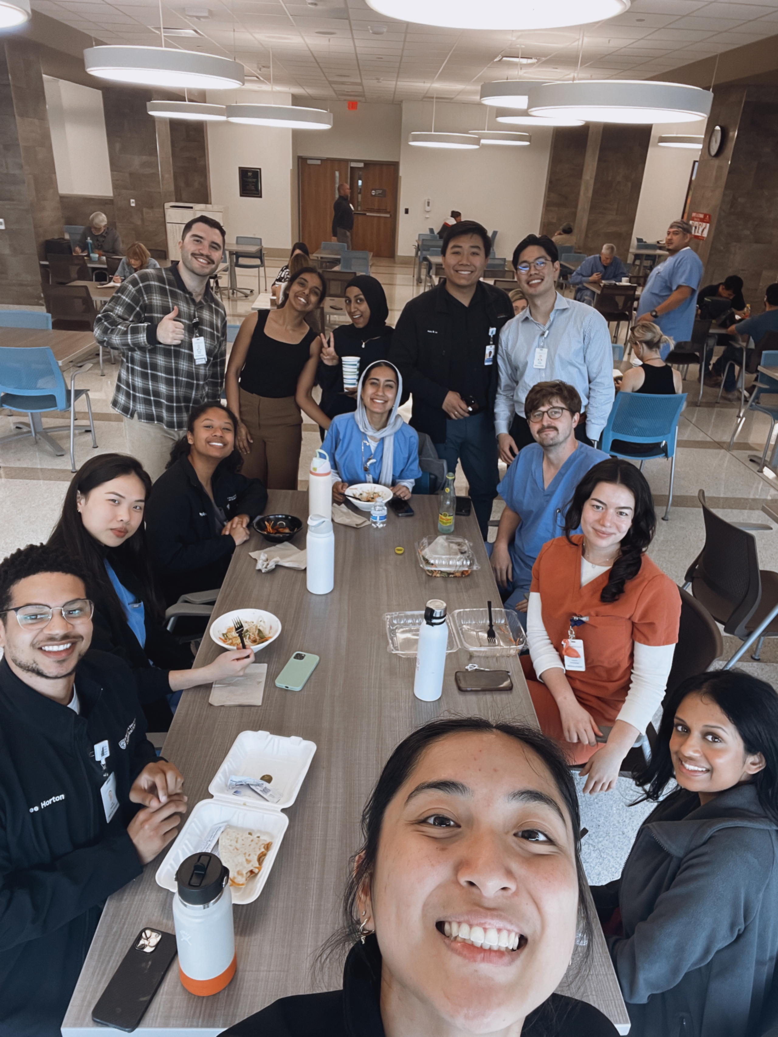 A Dell Medical School student takes a selfie in front of a dining hall table with several other students in the background.