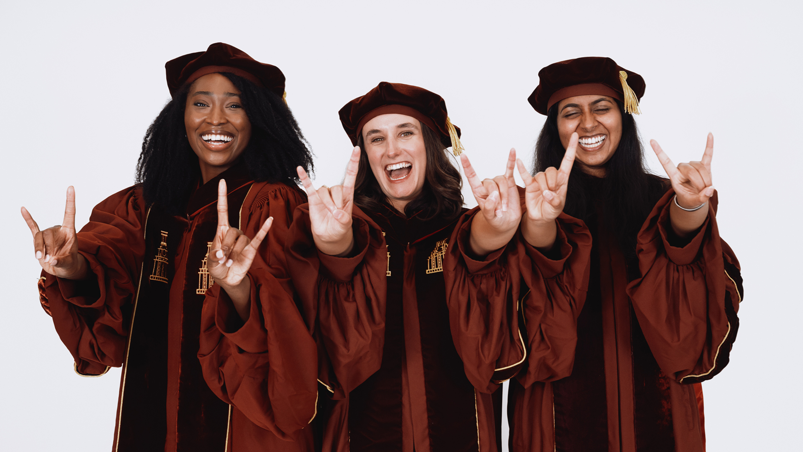 Three Dell Med students in graduation regalia doing the "Hook 'Em" gesture.