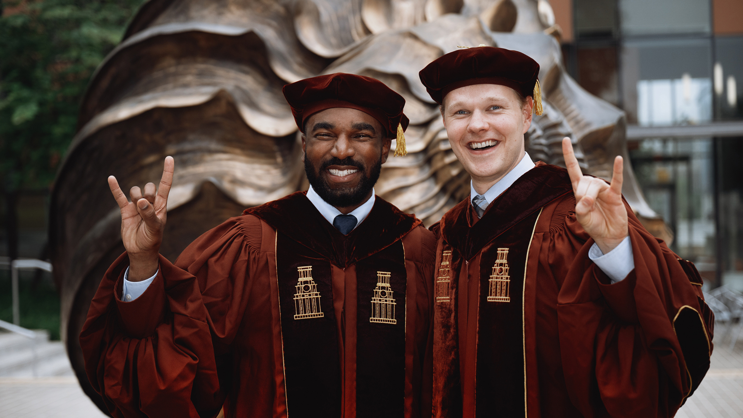 Cortney Matthews and Joshua Witkop stand together for a portrait outside the Dell Medical School campus. Both are wearing graduation regalia and are doing the Hook 'Em gesture.
