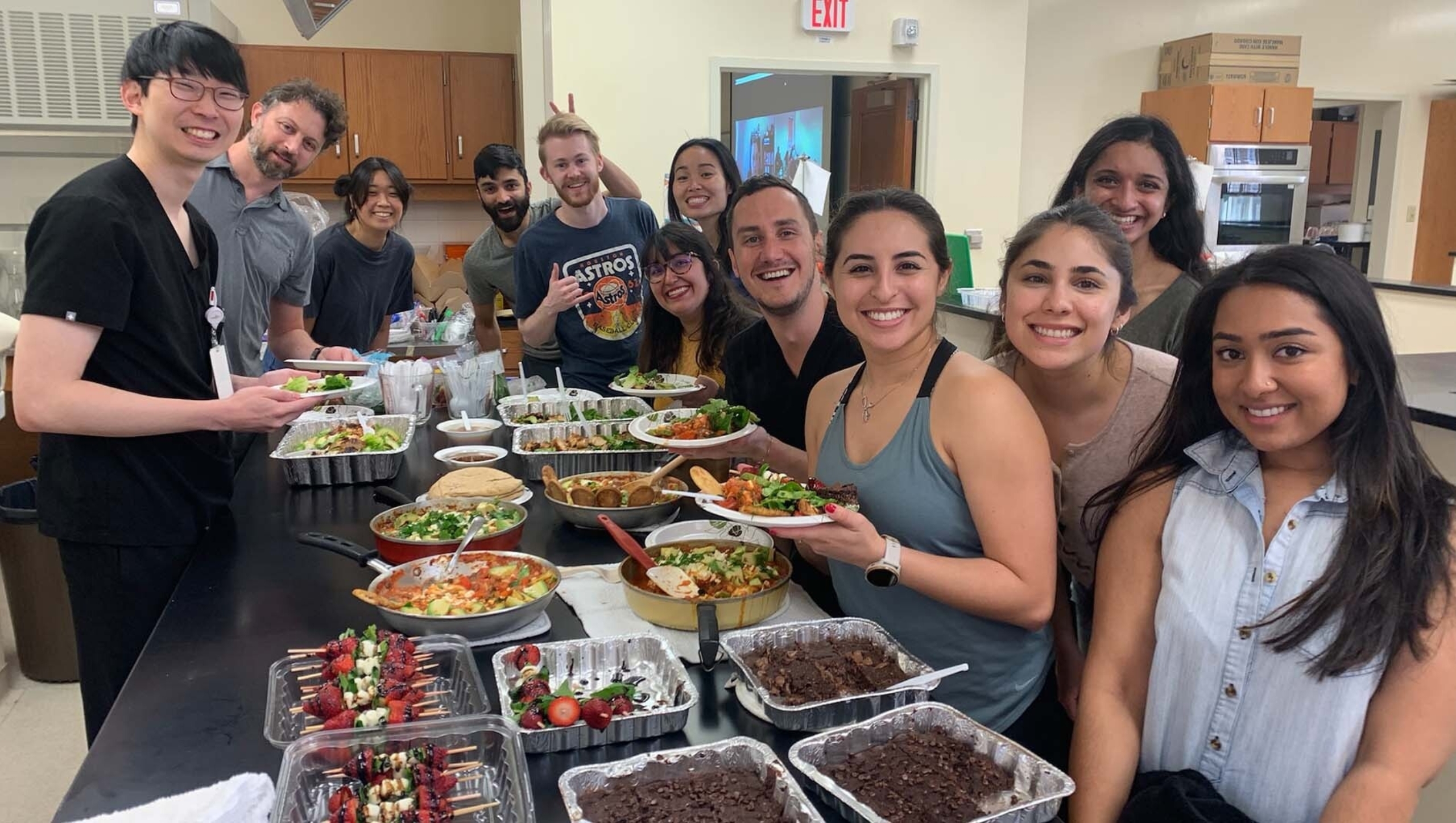 Pediatric residents and fellows gather around an assortment of food in a classroom kitchen for a group portrait.