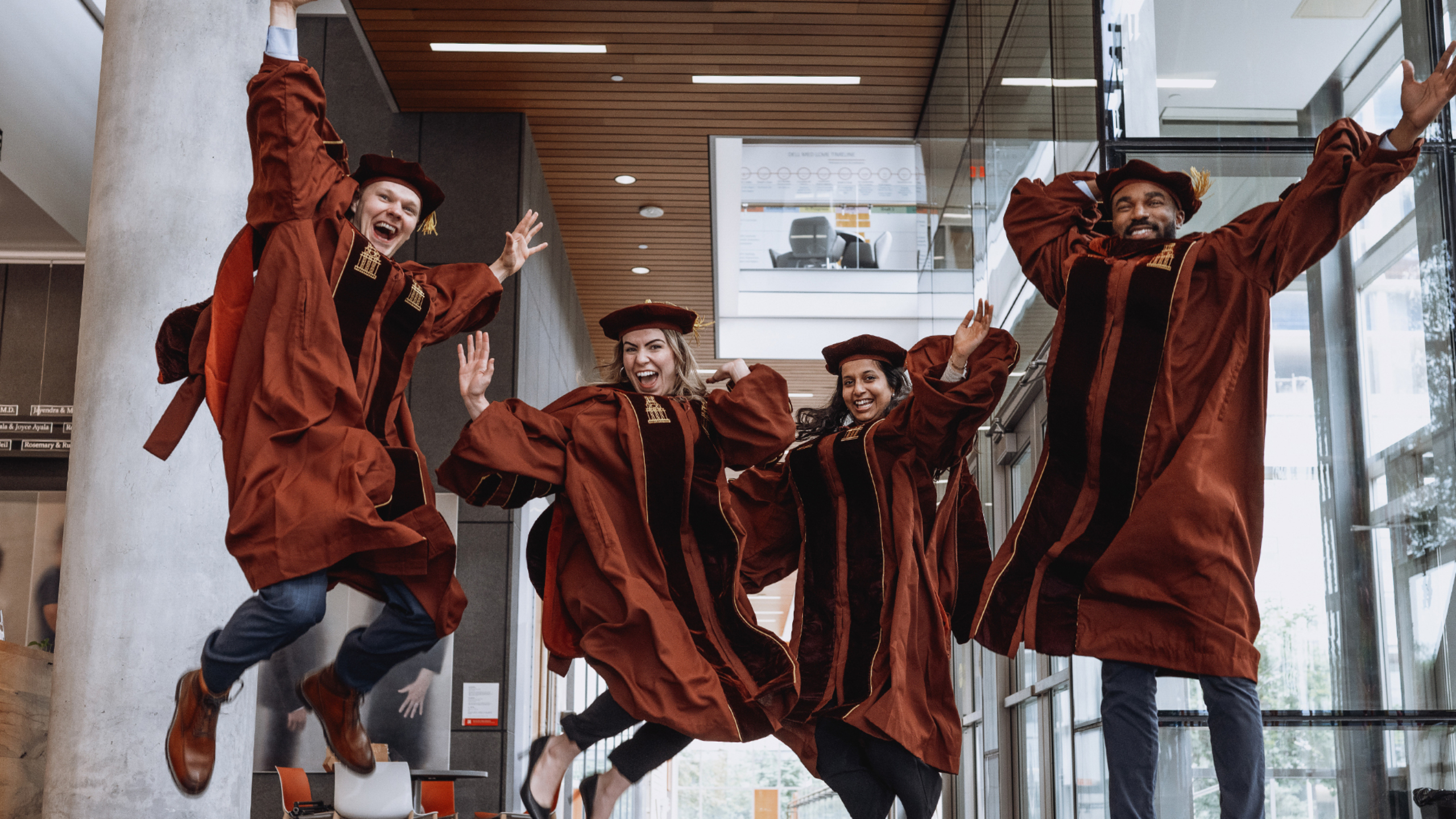 Four students from Dell Med's Class of 2024 — each wearing graduation regalia — jump for a celebratory portrait inside the Dell Med campus.