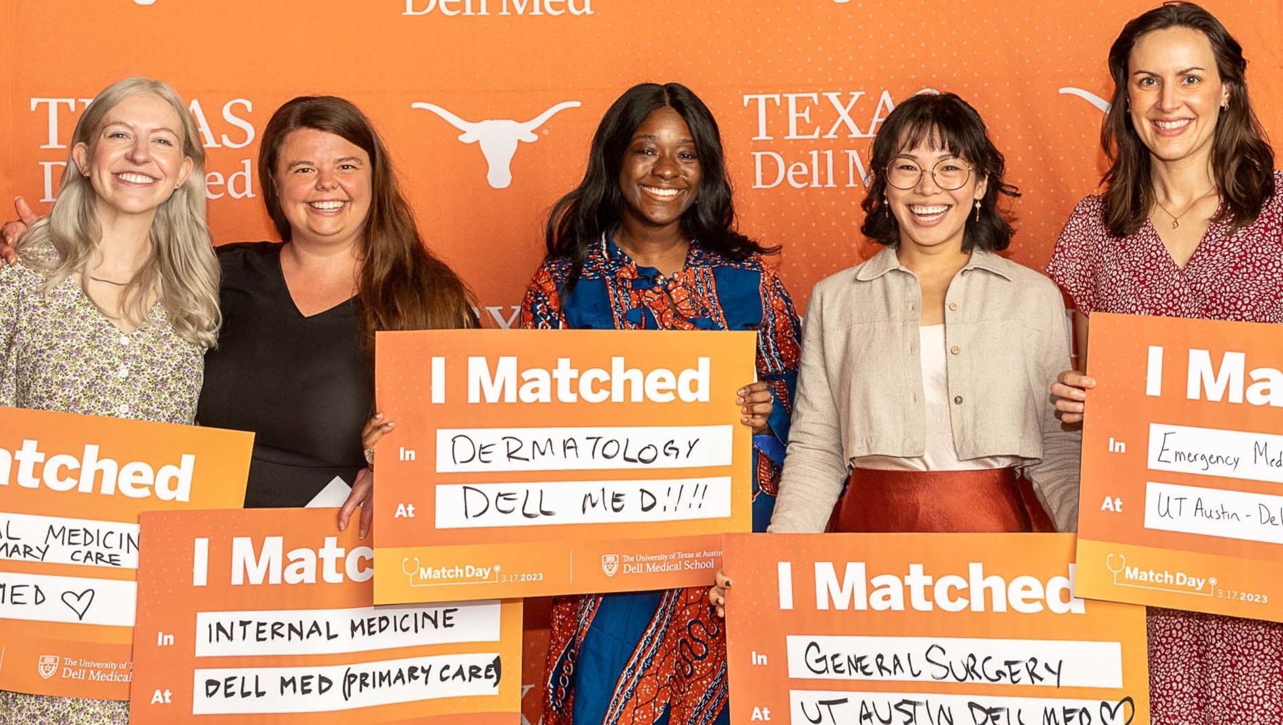Five women stand in front of a Dell Med backdrop holding Match day signs that indicate their respective residency program.