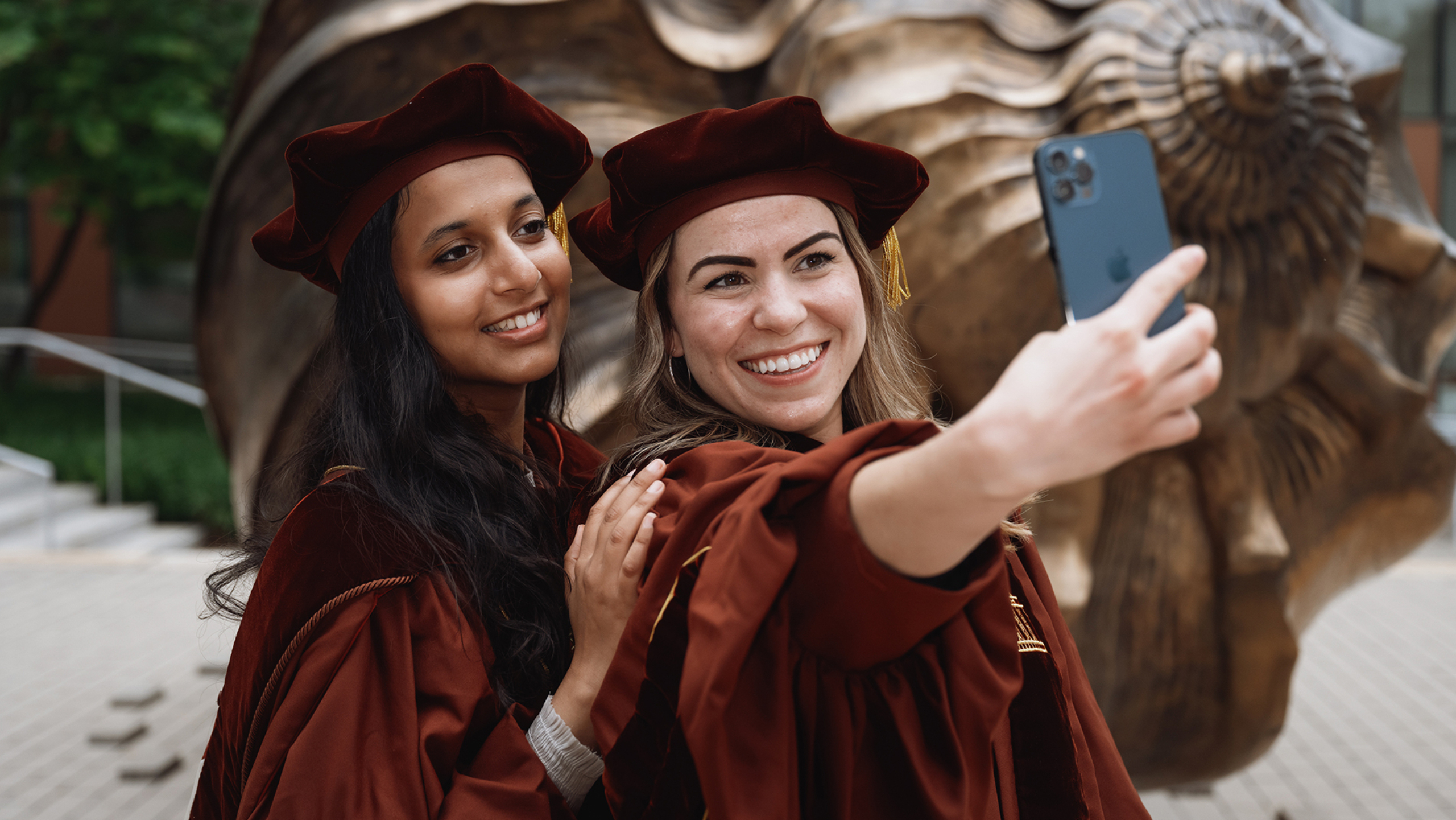 Nikita Choudhary stands behind Erin Kovar, who is holding out her phone for a selfie of the the two of them. Both Nikita and Erin are wearing graduation regalia, and in the background is an art sculpture that resides outside the Dell Med campus.