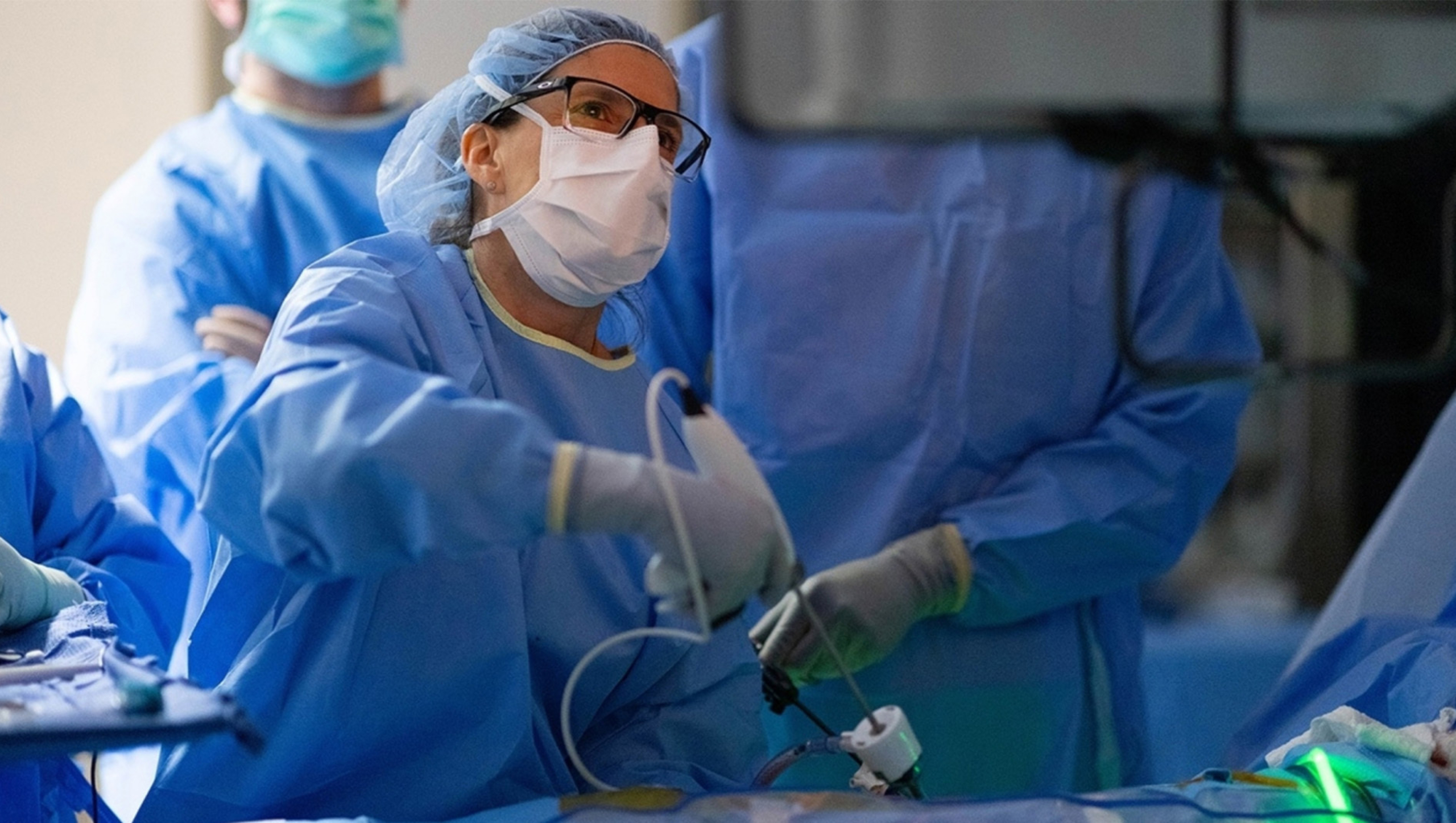 Nicole Turgeon, wearing blue medical scrubs, performs a procedure in an operating room. A number of surgeons in the background look on.