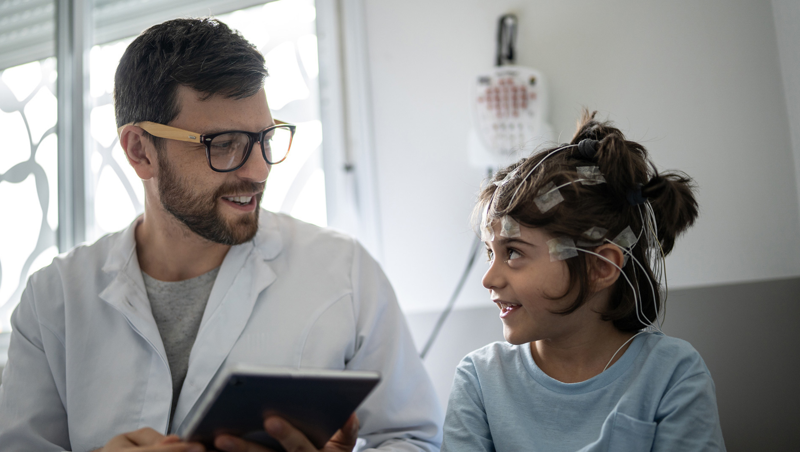 A male doctor looks at a reading on a tablet while a girl undergoes an EEG.