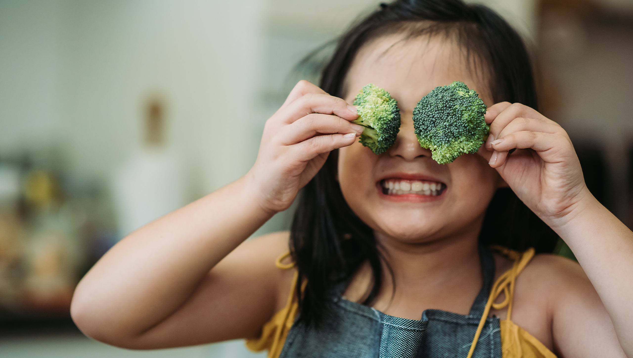 A young girl holds a piece of broccoli over each of her eyes.