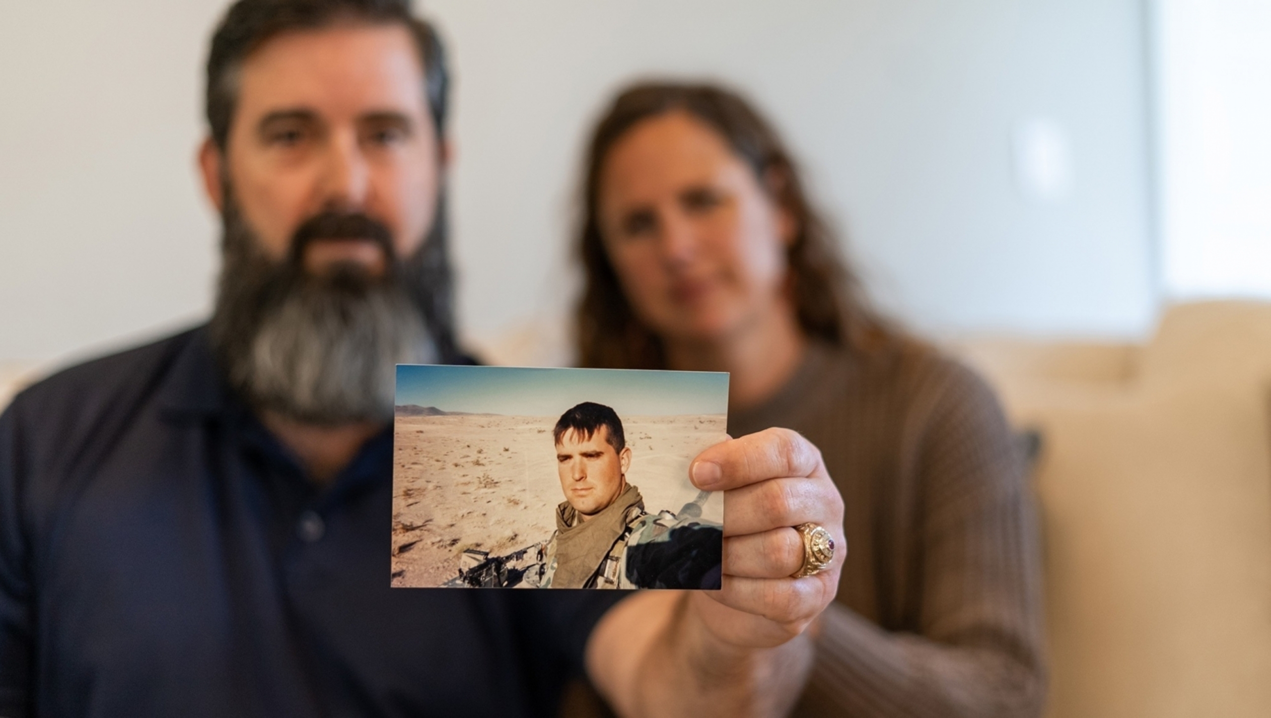 A man sitting beside his wife holds a photo of himself in military fatigues.