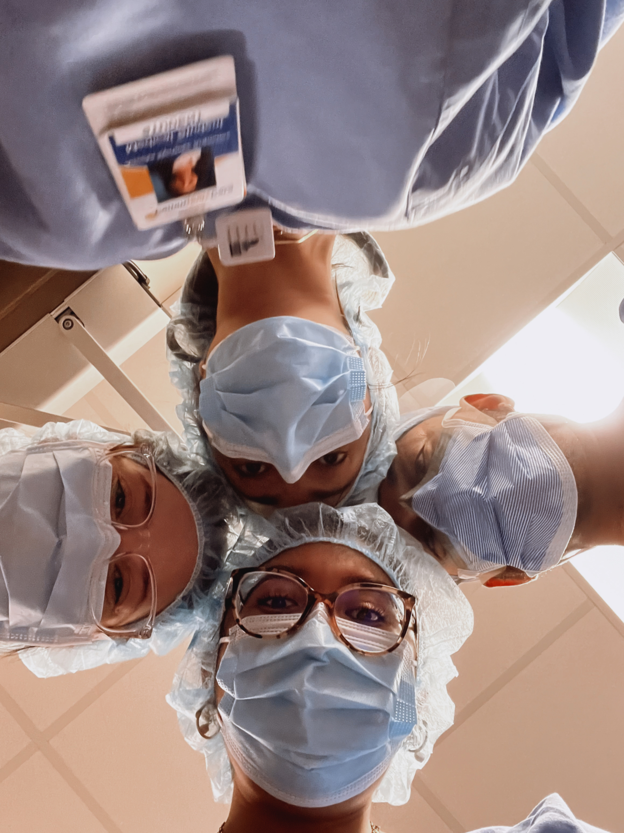 Four Dell Medical School students, wearing scrubs and medical masks, look down at a camera below them for a photo.