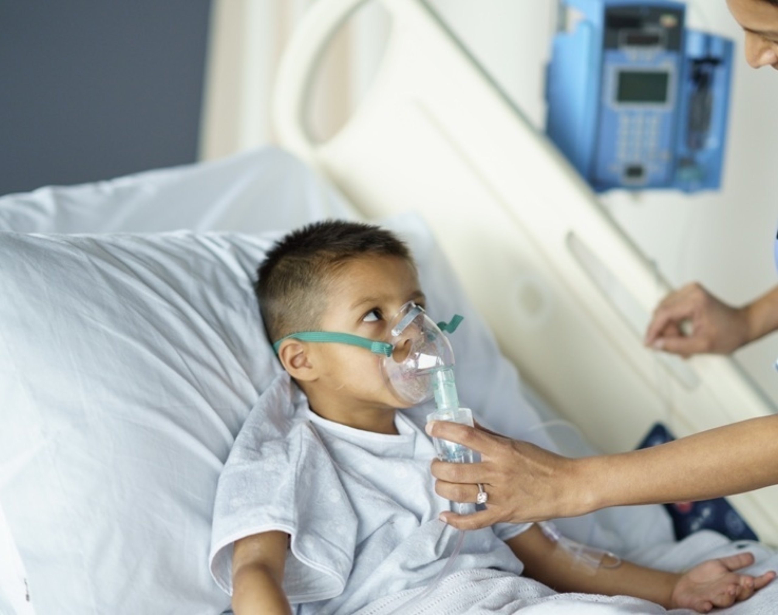 Young child receiving asthma treatment with a nebulizer mask in a hospital bed, assisted by a healthcare provider.